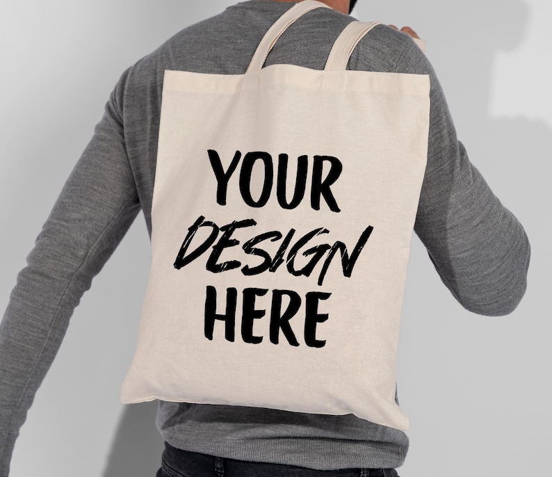 K&ouml;nnte beinhalten: Eine cremefarbene Tragetasche mit den Worten "YOUR DESIGN HERE" in fetter schwarzer Schrift. Die Tasche hat zwei Tr&auml;ger und wird &uuml;ber der Schulter getragen. Die Tasche ist aus einem robusten Stoff gefertigt.