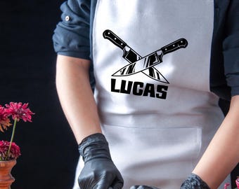 May include: White apron with a black design of two crossed knives and the name "LUCAS" printed below. A person is wearing the apron and holding a knife.