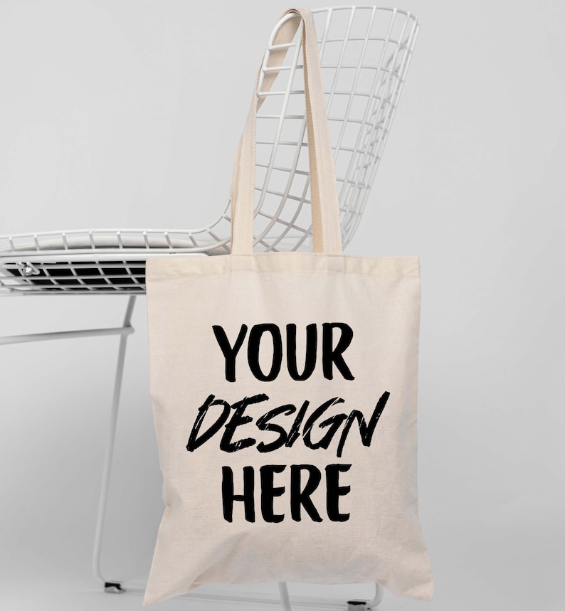 K&ouml;nnte beinhalten: Beige Tragetasche mit dem Text "YOUR DESIGN HERE" in Schwarz, h&auml;ngt an einem wei&szlig;en Drahtstuhl. Die Tasche hat lange Henkel und ein schlichtes Design, geeignet zum Tragen von Gegenst&auml;nden.