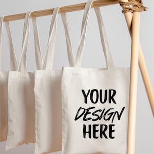 K&ouml;nnte beinhalten: Mehrere cremefarbene Tragetaschen h&auml;ngen an einer Holzstange. Die Taschen haben lange Henkel und eine rechteckige Form. Auf einer Tasche steht in schwarzer Schrift "YOUR DESIGN HERE". Die Stange ist aus hellem Holz gefertigt.