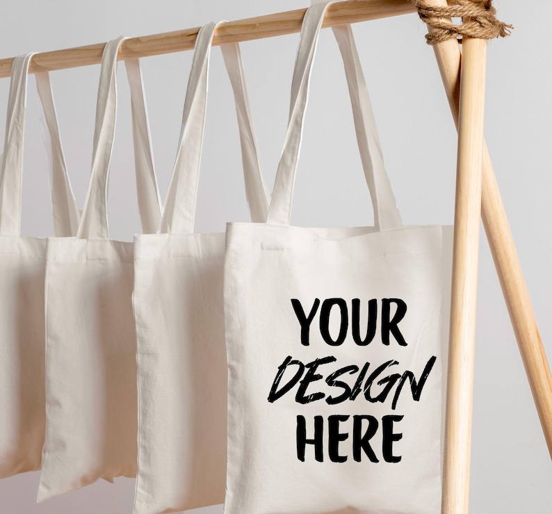 K&ouml;nnte beinhalten: Mehrere cremefarbene Tragetaschen h&auml;ngen an einer Holzstange. Die Taschen haben lange Henkel und eine rechteckige Form. Auf einer Tasche steht in schwarzer Schrift "YOUR DESIGN HERE". Die Stange ist aus hellem Holz gefertigt.