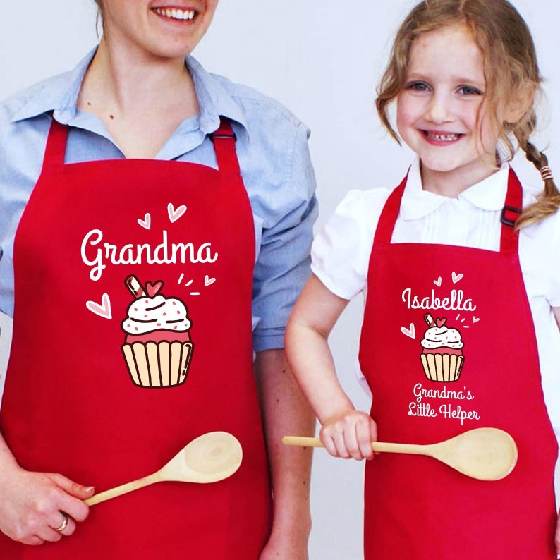 May include: Two red aprons with white text and cupcake designs. One apron says "Grandma" and the other says "Isabella Grandma's Little Helper". Both aprons have a wooden spoon.