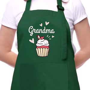 May include: A dark green apron with the word "Grandma" in white script, accompanied by a cupcake graphic. The cupcake has pink and white frosting, a red heart, and a brown wrapper. The apron has a pocket and adjustable neck strap.