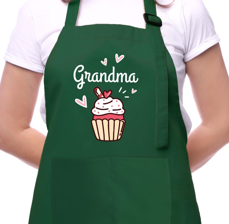 May include: A dark green apron with the word "Grandma" in white script, accompanied by a cupcake graphic. The cupcake has pink and white frosting, a red heart, and a brown wrapper. The apron has a pocket and adjustable neck strap.