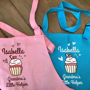 May include: Two aprons, one pink and one blue, with the name "Isabella" and a cupcake graphic. The text "Grandma's Little Helper" is below the cupcake. The aprons have adjustable straps.