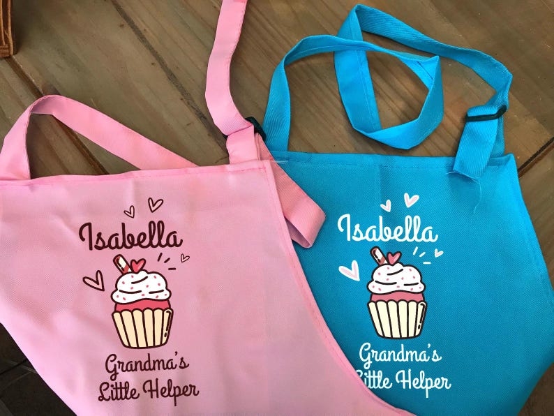May include: Two aprons, one pink and one blue, with the name "Isabella" and a cupcake graphic. The text "Grandma's Little Helper" is below the cupcake. The aprons have adjustable straps.