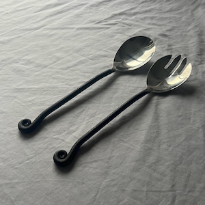 Puede incluir: Un par de utensilios para servir, una cuchara y un tenedor, con cabezas plateadas brillantes y mangos negros de estilo forjado. Los mangos tienen un remolino decorativo en el extremo. Para servir ensaladas u otros platos.
