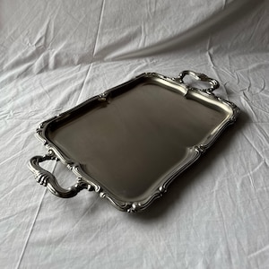 Può includere: Un vassoio rettangolare argentato con manici ornati e un bordo decorativo. Il vassoio ha una superficie riflettente ed è probabilmente in metallo. Le maniglie sono curve.