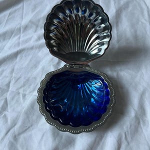 Peut inclure: Une boîte à bijoux en forme de coquillage, argentée et articulée, avec un intérieur bleu vif. La boîte est ouverte, révélant le design détaillé et nervuré de la coquille et la couleur bleue brillante à l'intérieur. Les bords sont festonnés avec une bordure perlée.