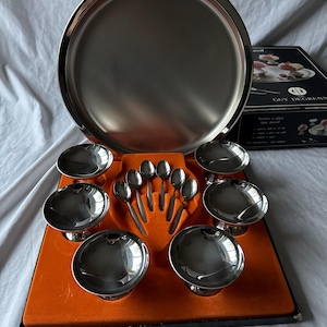 Puede incluir: Un juego de servir vintage de acero inoxidable Guy Degrenne. El juego incluye una bandeja redonda grande, ocho cuencos pequeños y ocho cucharas, todos con una superficie brillante y reflectante. Los cuencos están sobre pedestales. El juego se exhibe en un inserto de terciopelo naranja.