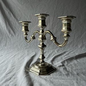 Può includere: Un candelabro color argento con tre bracci, ciascuno con un portacandele. Il candelabro ha una base decorativa e bracci curvi. Il metallo ha una finitura lucida, che riflette la luce. È un oggetto decorativo.