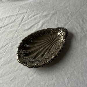 Op de afbeelding: Een zilverkleurige, schelpvormige schaal met een gedetailleerde, sierlijke rand. De schaal heeft een geribbelde binnenkant en een decoratief ontwerp aan één uiteinde. De schaal is waarschijnlijk van metaal en ligt op een wit oppervlak.