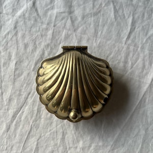 May include: A gold-colored, seashell-shaped trinket box. The box is made of metal and has a hinged lid. The shell design features detailed ridges and a small, round knob at the base. The box is closed and sits on a white, textured surface.