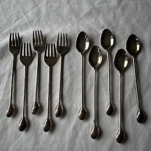 May include: An assortment of silver-coloured forks and spoons displayed on a light-coloured surface. The forks have three tines and a textured handle. The spoons have a rounded bowl and a matching handle design. These utensils are likely for serving or eating.
