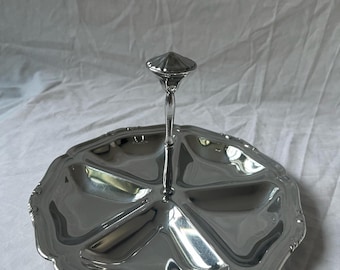 May include: A silver-colored serving tray with a handle. The tray is divided into six sections and has a scalloped edge. The handle is a slender stem topped with a decorative, conical finial. The tray is reflective and appears to be made of metal.