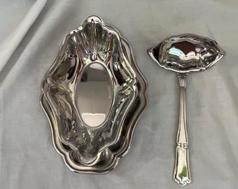 Puede incluir: Un juego de salsera y cuchara plateadas. La salsera tiene un borde festoneado y un interior ovalado. La cuchara tiene un mango decorativo y un cuenco en forma de concha. El juego es probablemente de metal.