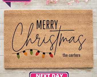 May include: A rectangular doormat with the words "MERRY Christmas" in a stylish script, with a string of colorful lights. The text "the carters" is below the word Christmas. The mat is a natural tan color.