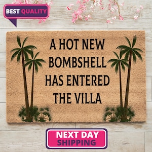 May include: A rectangular tan coir doormat featuring black text that reads A HOT NEW BOMBSHELL HAS ENTERED THE VILLA. The design includes two green palm tree illustrations, one on the left side and one on the right side of the central text.