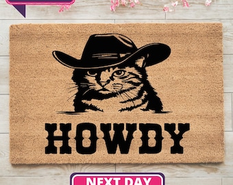 Może przedstawiać: Brązowa mata kokosowa z czarną sylwetką kota w kowbojskim kapeluszu. Tekst "HOWDY" jest nadrukowany na czarno pod kotem.