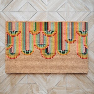 May include: A doormat with a colorful rainbow design. The rainbow arches are made of stripes in shades of red, orange, yellow, green, blue, indigo, and violet. The doormat is made of coir and has a natural brown color.