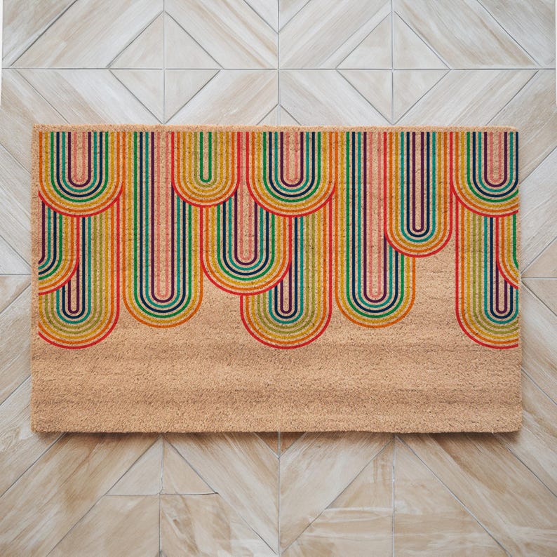 May include: A doormat with a colorful rainbow design. The rainbow arches are made of stripes in shades of red, orange, yellow, green, blue, indigo, and violet. The doormat is made of coir and has a natural brown color.