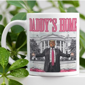 May include: White ceramic mug with a pink "Daddy's Home" text and a cartoon image of a man in a suit standing in front of the White House.