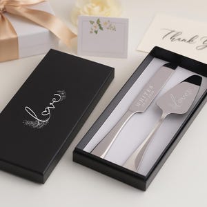 May include: A wedding cake knife and server set in a black box. The server has the word "love" engraved on it. The box is next to a gift box with a gold ribbon and a card that says "Thank You".