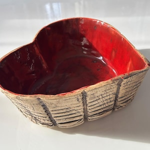 May include: A heart-shaped ceramic bowl with a red glaze inside and a textured, off-white exterior.