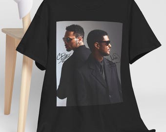 May include: Black t-shirt featuring a photo of two men wearing sunglasses and black suits. The image has signatures on it. A white baseball cap rests on a wooden chair in the background.
