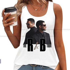 Könnte beinhalten: Weißes Tanktop mit einer Grafik von zwei Männern in schwarzen Anzügen und Sonnenbrillen, mit dem Text "THE R&B TOUR" in Schwarz. Das Model trägt eine blaue Jeans und hält eine Kaffeetasse.