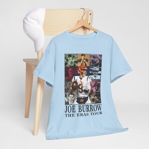 Pu&ograve; includere: Maglietta blu chiaro con un collage di foto di Joe Burrow, un giocatore di football americano, in diverse pose e uniformi. Il testo "Joe Burrow The Eras Tour" &egrave; stampato sotto il collage.