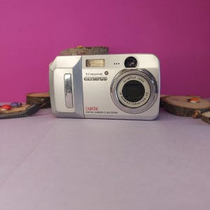 May include: Silver Olympus Camedia digital camera with a zoom lens. The camera has a 5.0 megapixel resolution and features a built-in flash. The camera is on a light purple surface.