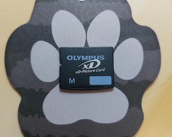 May include: Black Olympus xD-Picture Card with white text and a blue accent. The card is labeled with the Olympus logo and "xD-Picture Card". The card is on a gray paw-shaped background.
