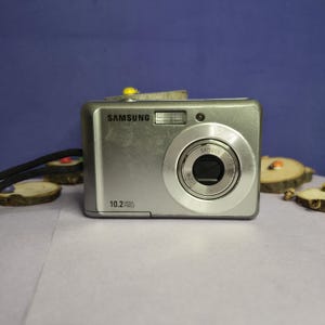 May include: A silver Samsung digital camera with a prominent lens and flash. The camera has the text "SAMSUNG" and "10.2 MEGA PIXELS" printed on it. The camera is on a light purple surface.
