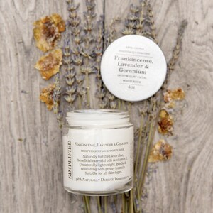 Könnte beinhalten: Ein weißes Glas mit Gesichtscreme mit einem schwarzen Etikett, auf dem "SIMPLIFIED FRANKINCENSE, LAVENDER & GERANIUM LIGHTWEIGHT FACIAL MOISTURIZER" und "98% NATURALLY DERIVED INGREDIENTS" steht. Das Glas steht auf einer Holzoberfläche mit Lavendel- und Bernsteinblumen.