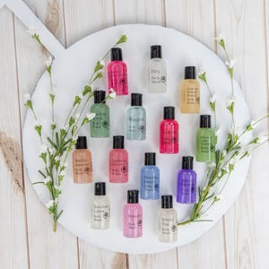 Puede incluir: Una bandeja circular blanca con una variedad de pequeños frascos de gel de baño. Los frascos están dispuestos en círculo y están etiquetados con diferentes aromas, incluyendo "Vanilla Jasmine", "Coconut & Lime", "Sunday French", "Bamboo Lotus", "White Tea", "Fifty", "Rugged", "Parisian Mint", "Cream Milk & Honey", "Citrus Verbena", "Lemon Lavender", "Rosemary" y "Ocean Mist".