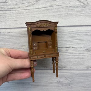 Puede incluir: Mini escritorio marrón con detalles dorados y pequeños cajones. El escritorio tiene una alacena con la parte superior curvada y está sostenido por patas delgadas. Un mueble pequeño y detallado.