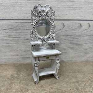 May include: A miniature white vanity with an oval mirror and swan-shaped accents. The vanity has a small drawer and a lower shelf. The mirror is framed with ornate detailing. The overall design is vintage and decorative.