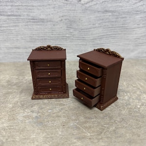 Puede incluir: Dos cómodas de madera marrón en miniatura con herrajes dorados. Una cómoda tiene todos los cajones cerrados, mientras que la otra tiene cuatro cajones abiertos. Ambas tienen detalles ornamentados en la parte superior e inferior, sobre fondo gris claro.
