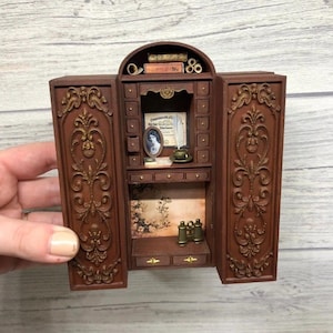May include: A detailed miniature wooden cabinet with decorative carvings and gold detailing. Inside are tiny drawers, a framed portrait, books, and miniature binoculars. The cabinet doors are open, showcasing the interior and floral wallpaper.