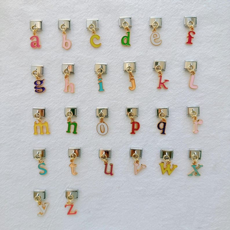 Pu&ograve; includere: Una collezione di ciondoli alfabetici colorati, ciascuno con una chiusura argentata e dettagli dorati. Le lettere sono in vari colori, tra cui rosa, verde, giallo e blu. I ciondoli sono disposti in file e colonne.