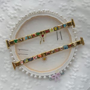 May include: Two gold-tone modular link bracelets featuring various colorful enamel charms, including a red heart, a daisy, a bow, and a yin-yang symbol. The bracelets are displayed with four small metal connector pins on a white circular frame.