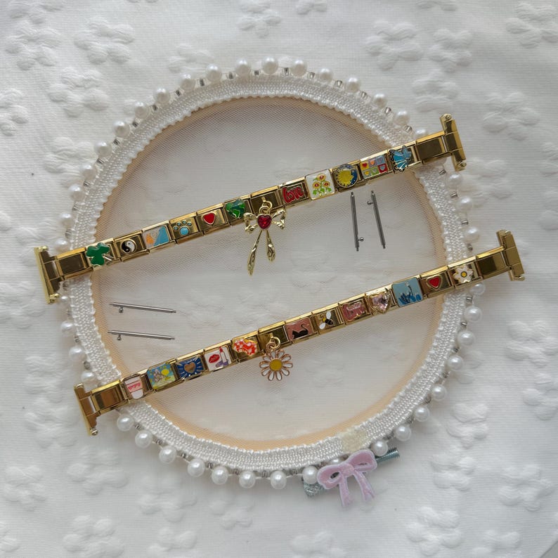 May include: Two gold-tone modular link bracelets featuring various colorful enamel charms, including a red heart, a daisy, a bow, and a yin-yang symbol. The bracelets are displayed with four small metal connector pins on a white circular frame.