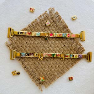 May include: Two gold charm bracelets with colourful enamel charms, including hearts, stars, and flowers. The bracelets are displayed on a textured brown surface with scattered charms. One bracelet has a green charm and the other has the letters "M" and "J".