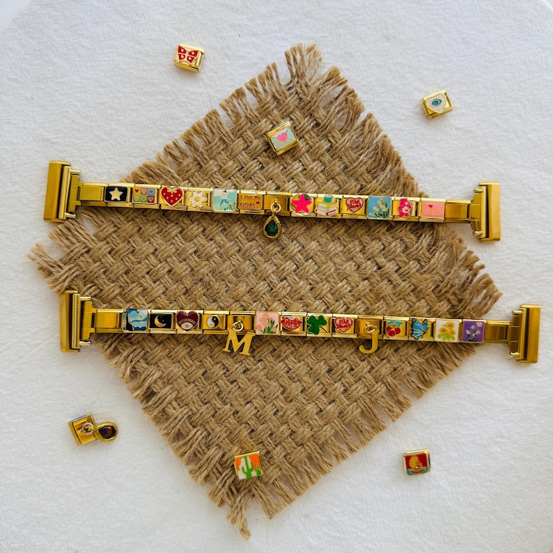 May include: Two gold charm bracelets with colourful enamel charms, including hearts, stars, and flowers. The bracelets are displayed on a textured brown surface with scattered charms. One bracelet has a green charm and the other has the letters "M" and "J".