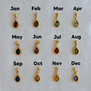 May include: Twelve gold-toned teardrop pendants, each featuring a different colored gemstone representing the months of the year, labeled Jan through Dec in black text above each charm, arranged in a grid on a white textured background.