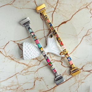 Pu&ograve; includere: Due braccialetti decorativi con ciondoli quadrati colorati. Un braccialetto &egrave; argentato, l'altro dorato. Ogni ciondolo presenta un design diverso, tra cui fiori, frutta e motivi astratti. I braccialetti sono esposti su un oggetto decorativo bianco su sfondo marmoreo.