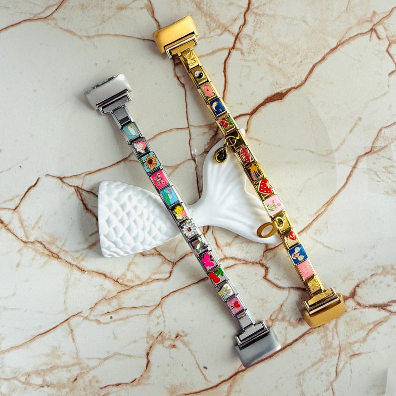 Pu&ograve; includere: Due braccialetti decorativi con ciondoli quadrati colorati. Un braccialetto &egrave; argentato, l'altro dorato. Ogni ciondolo presenta un design diverso, tra cui fiori, frutta e motivi astratti. I braccialetti sono esposti su un oggetto decorativo bianco su sfondo marmoreo.