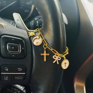 May include: A gold-toned chain bracelet featuring multiple charms including a star, a yin-yang symbol, a cross with a central stone, a small bow, and a circular pendant with a cross emblem. The bracelet is constructed with elongated oval links.