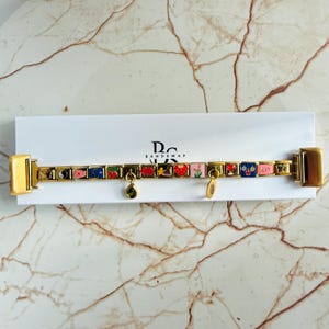 Puede incluir: Pulsera dorada con abalorios de esmalte de colores. Los abalorios incluyen una fresa, una flor y la palabra "sexy". La pulsera se muestra en una caja blanca con el texto "Bandswap".